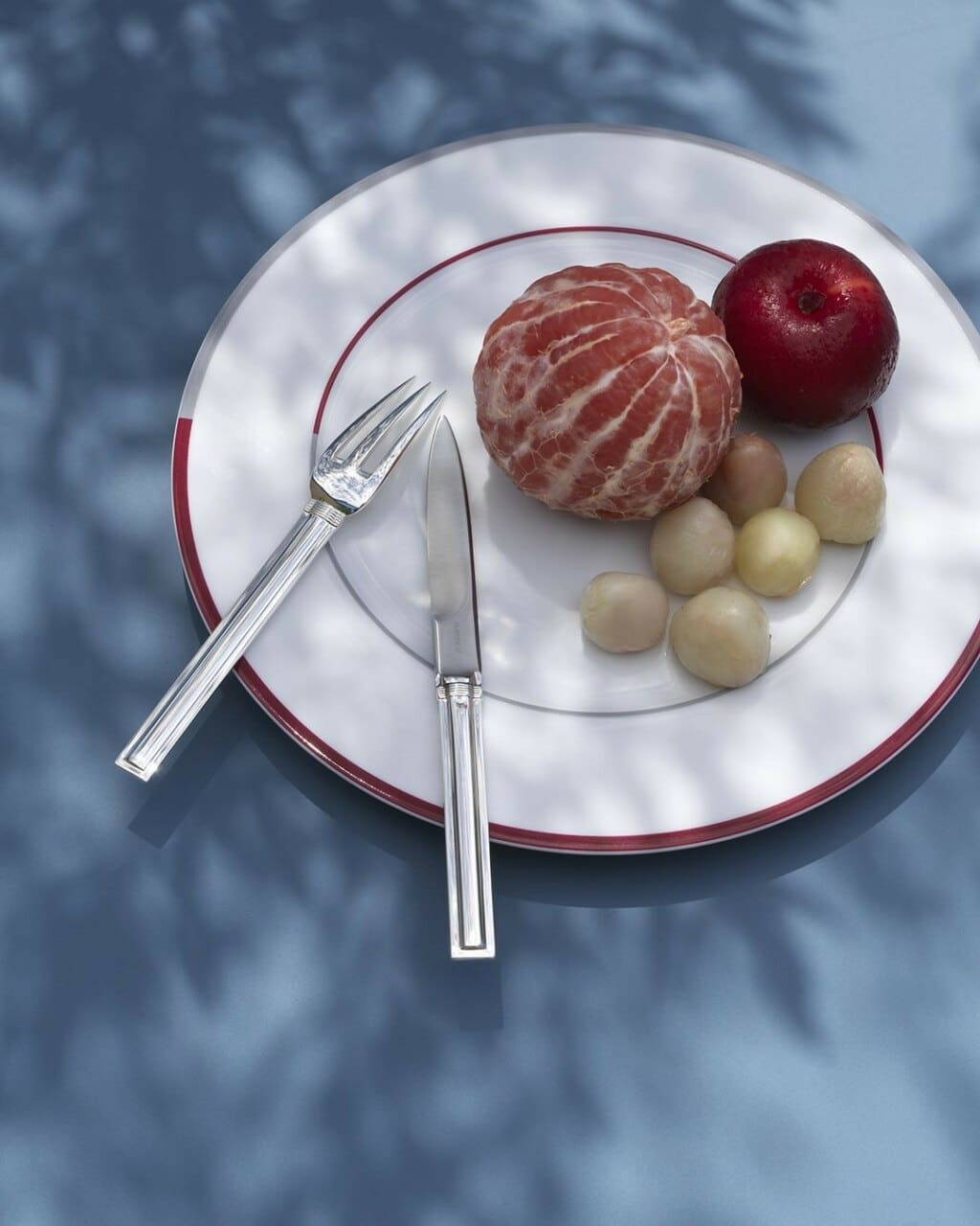 Puiforcat Cannes collection in sterling silver - set of table cutlery in an Initiales plate full of fruits under a shady tree on a grey tablecloth