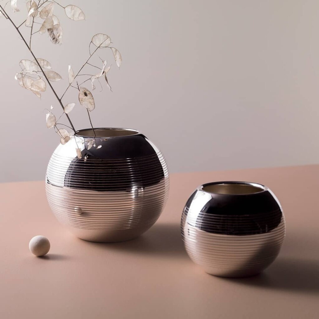 Vases en métal argenté de la collection Pétanque de Puiforcat mis en scène sur une table rose pale avec une branche de feuillage et un cochonnet