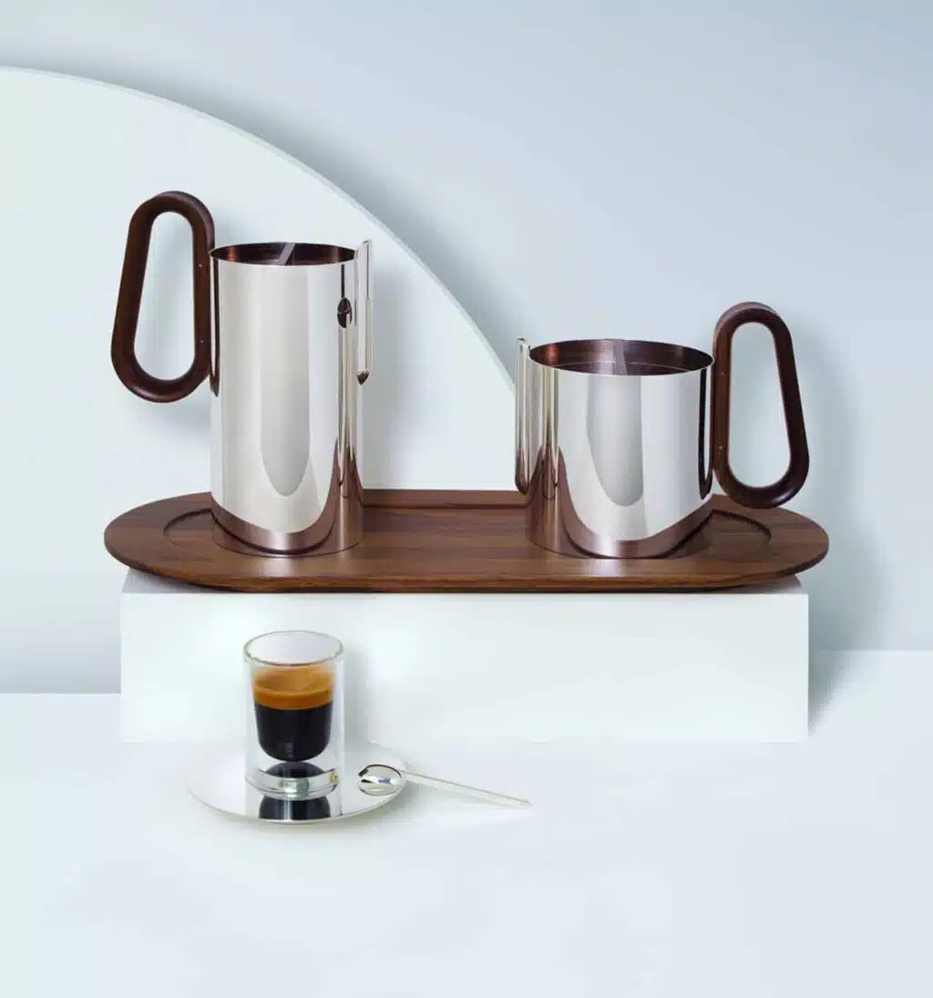 Mise en scène de la cafetière et de la théière en métal argenté sur un plateau en bois de la collection Phi sur un fond blanc et bleu