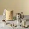 Tea pot, creamer, expresso and infusion cups and saucers photographed from side on a marble table with a big pastry and a cake shovel in front of a pale yellow wall