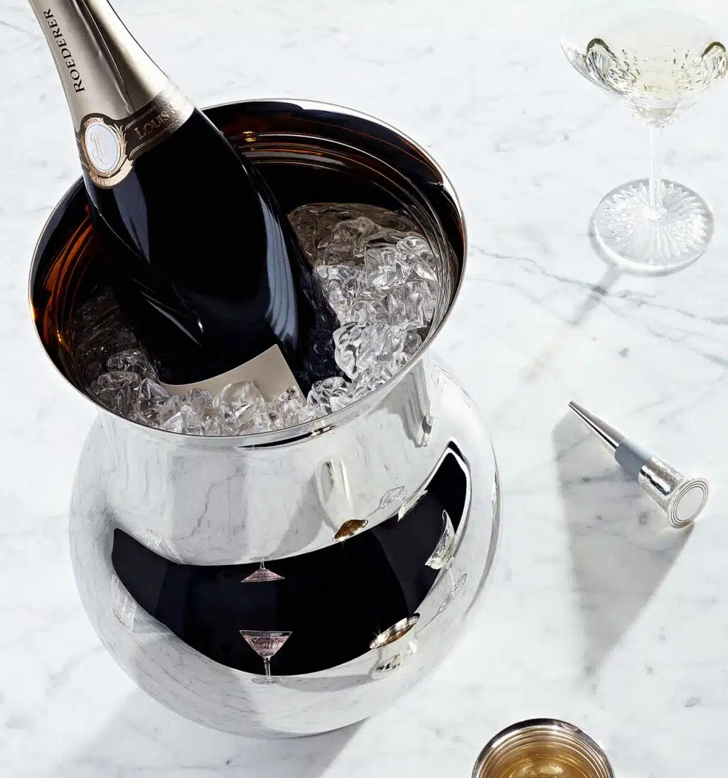 Seau à champagne en argent massif de la collection Phi rempli de glaçons et d'une bouteille de champagne, photographiés à côté d'un verre en cristal de la cristallerie Saint Louis, d'un bouchon de champagne en argent massif de Puiforcat et d'une timbale en argent massif de la même collection, sur une table en marbre