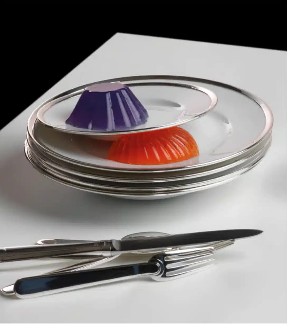 Pile d'assiettes de la collection Cercle d'orfèvre entremêlées de flan. La pile est posée à côté de couverts de différentes collections de Puiforcat sur une table blanche