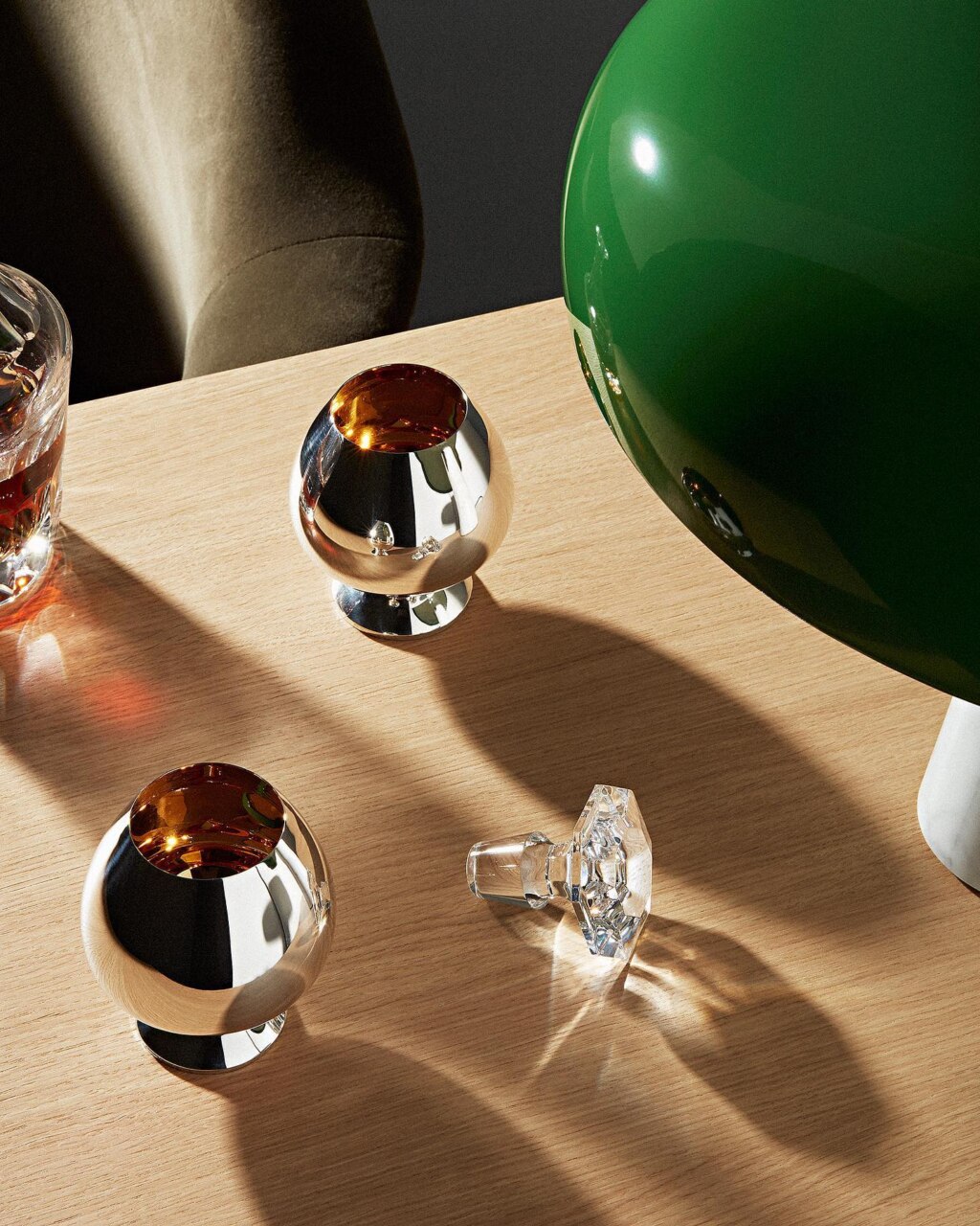 Cognac tumblers in sterling silver on a wooden table, photographed next to a green lamp and a bottle cap