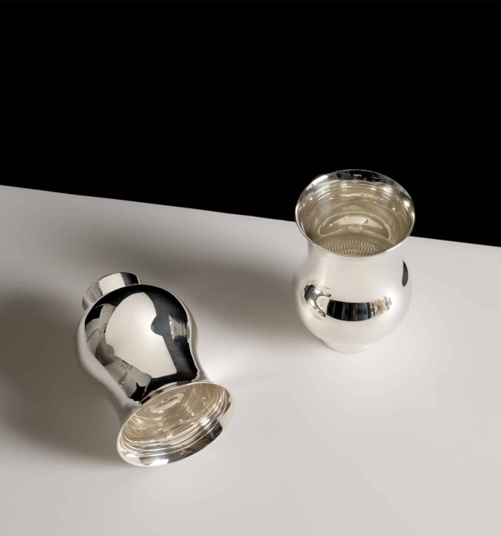 Two tumblers in sterling silver (one standing, the other lying on side) from Pour le Champagne collection, on a white table, in front of a black wall