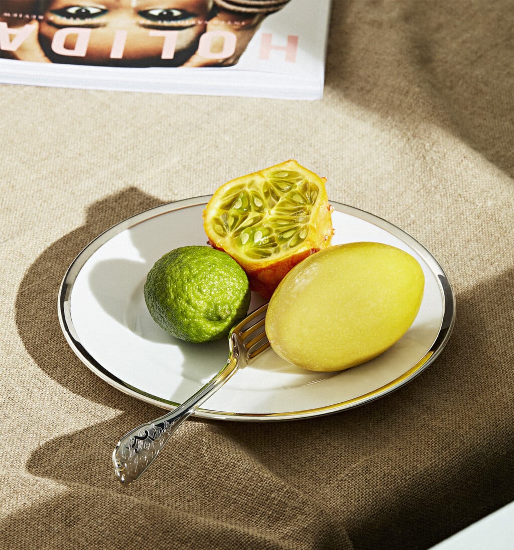 Assiette de la collection Cercle d'orfèvre présentant trois fruits et une fourchette de la collection Elysée de Puiforcat, posés sur une nappe beige à côté d'un magazine féminin