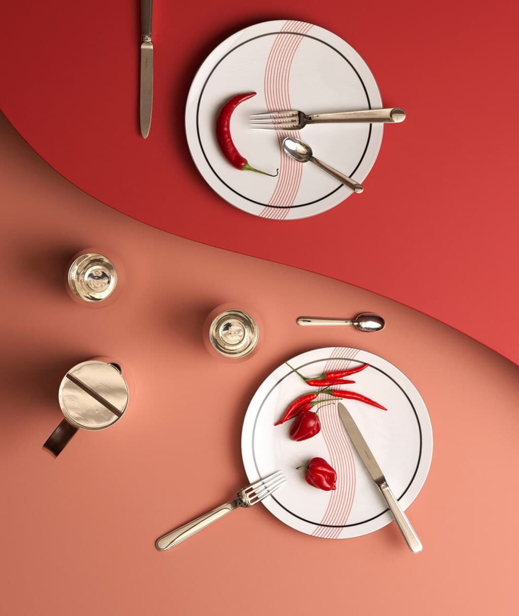 Porcelain plates from Initiales collection on a pink and red background, photographed with pieces of cutlery and table accessories from Phi collection