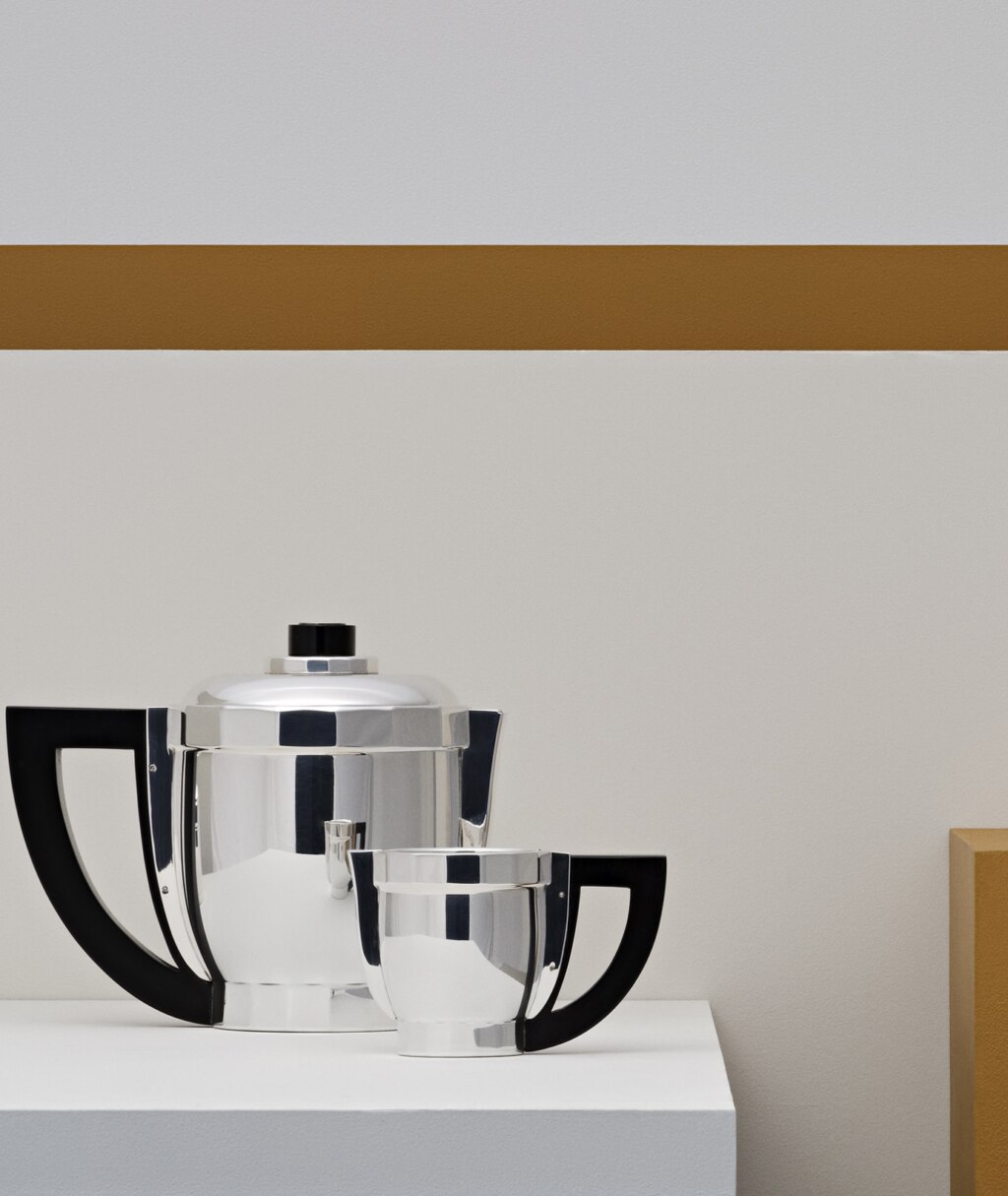 Coffee pot and creamer in silver plated from Loya Art Déco 1927 collection from Puiforcat pictured on a white stele in front of a white wall decorated with a mustard band and stele