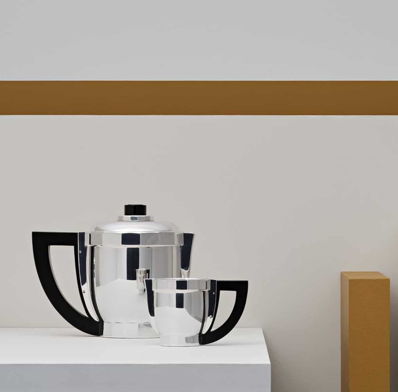 Coffee pot and creamer in silver plated from Loya Art Déco 1927 collection from Puiforcat pictured on a white stele in front of a white wall decorated with a mustard band and stele