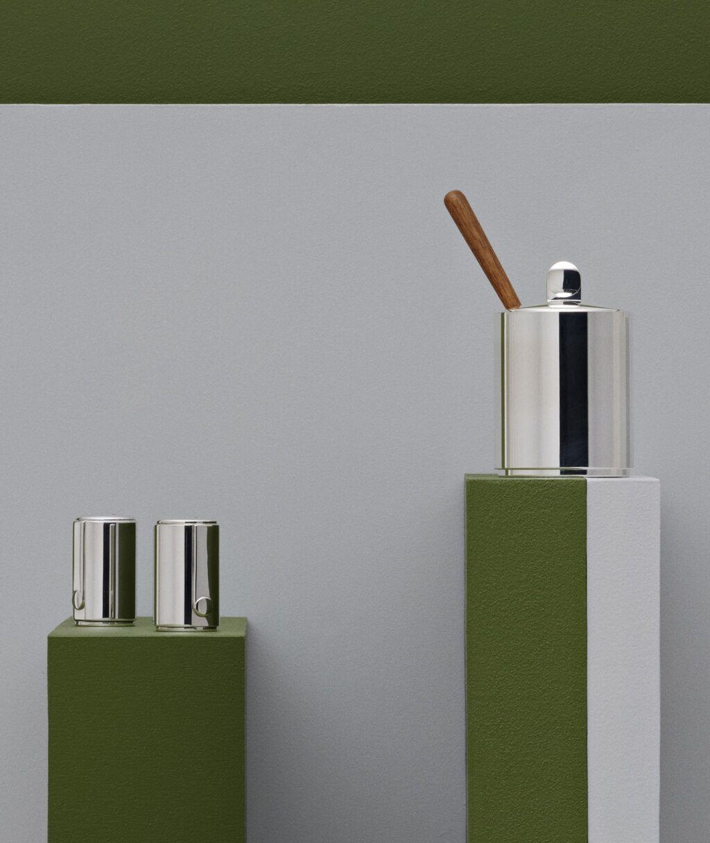 Salt and pepper mills and mustard pot in silver plated from Normandie collection from Puiforcat photographed on steles of different heights in front of a grey and green wall