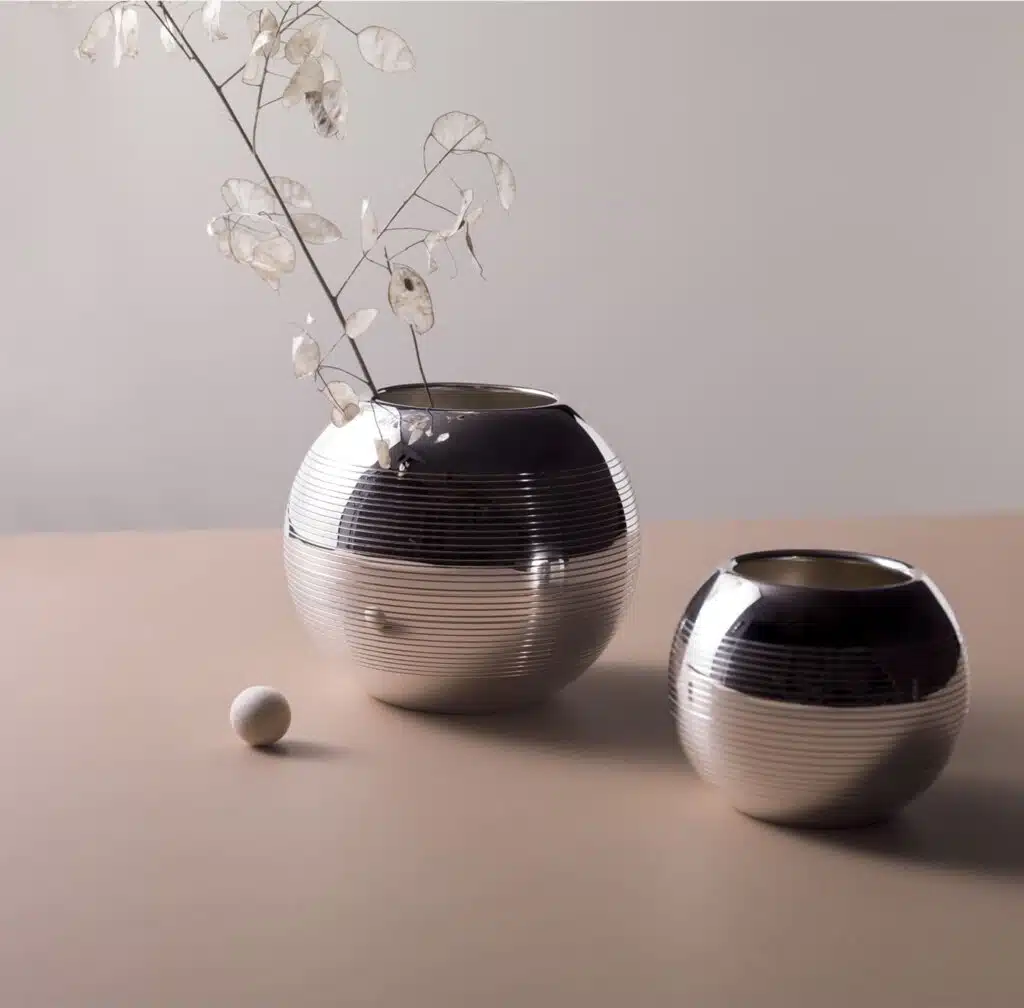 Vases in silver plated from Pétanque collection from Puiforcat photographed on a light pink table in front of a grey wall with a flower in the left vase, and a boule game starter in front