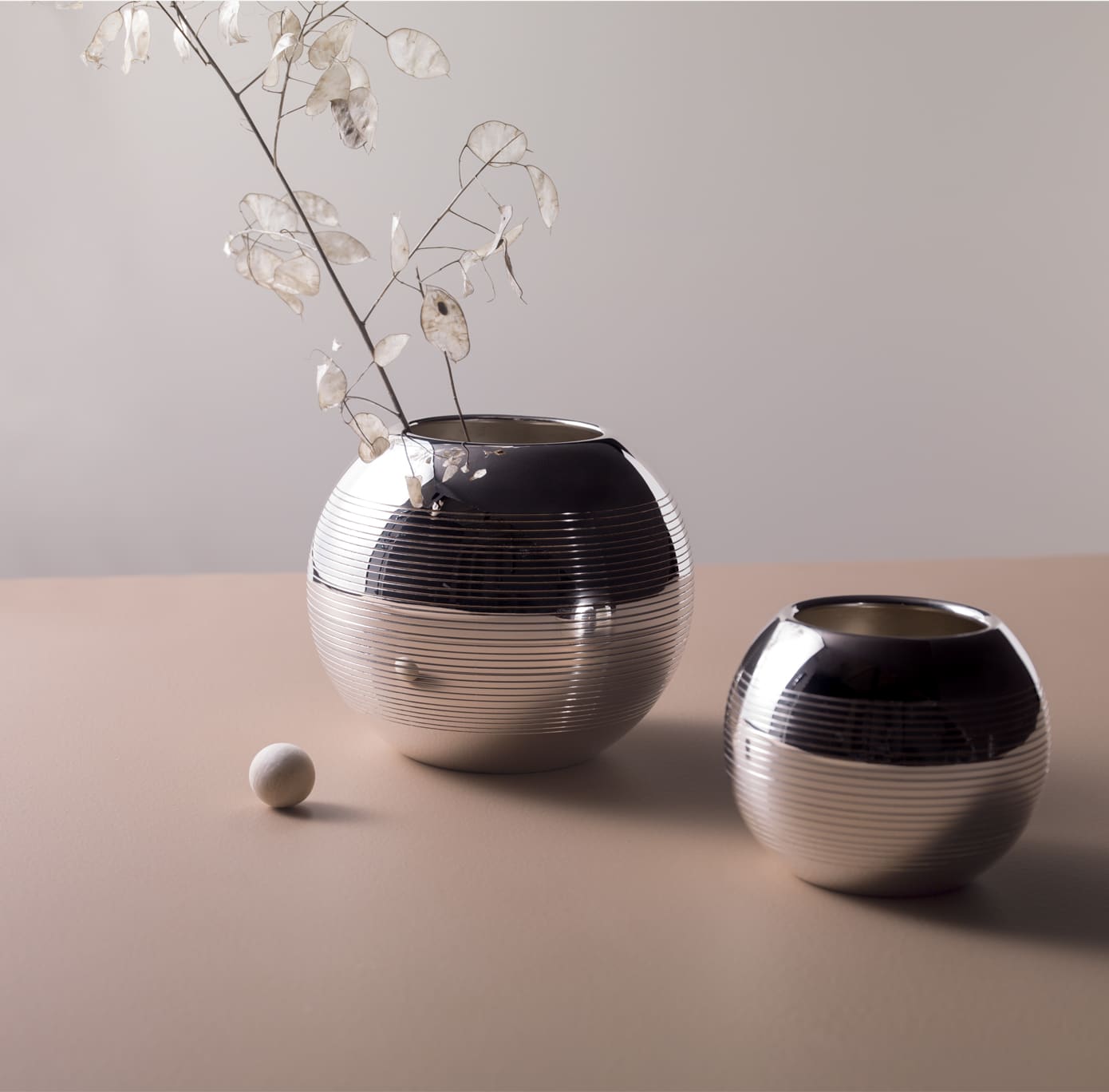 Vases in silver plated from Pétanque collection from Puiforcat photographed on a light pink table in front of a grey wall with a flower in the left vase, and a boule game starter in front