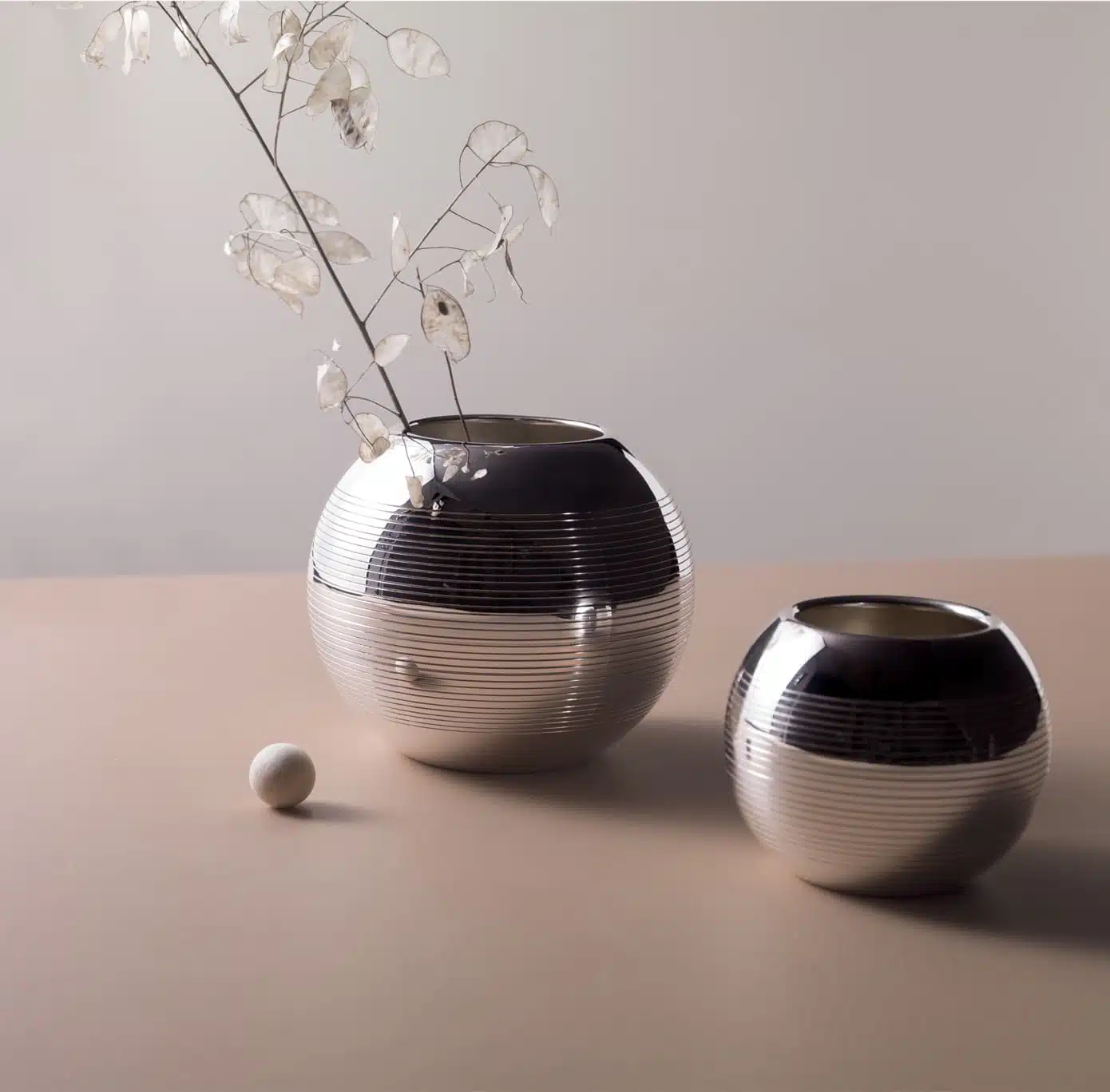 Vases en métal argenté de la collection Pétanque de Puiforcat mis en scène sur une table rose pale avec une branche de feuillage et un cochonnet