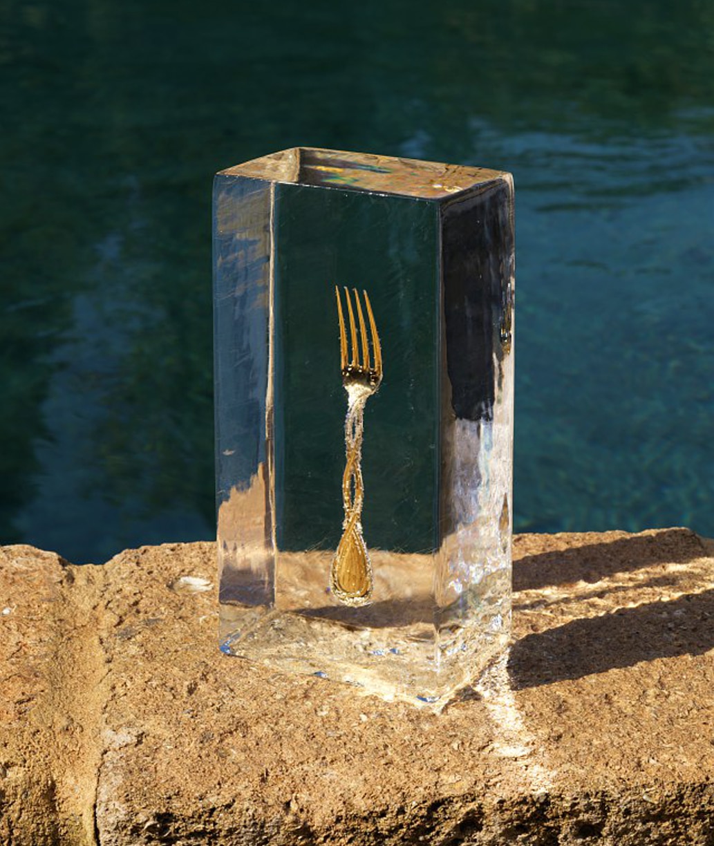 Fourchette de table en argent massif et finition or de la collection Royal de Puiforcat photographiée dans un bloc de glace, posé sur un rebord de pierre devant une rivière