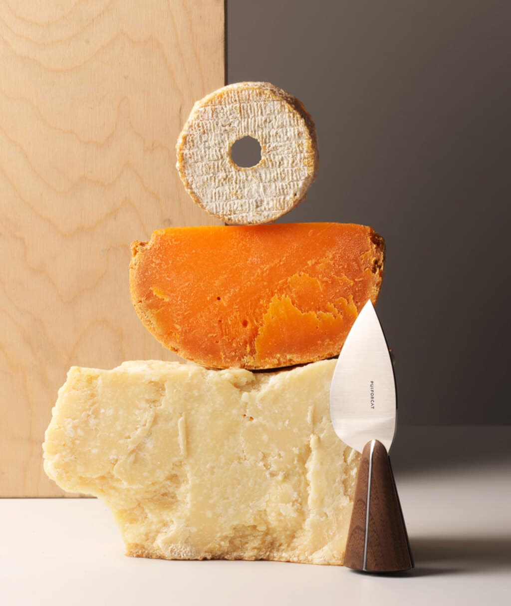 Couteau à fromage de la collection Couteaux d'orfèvre posé à côté d'une pile de trois fromages, photo prise sur un fond en bois