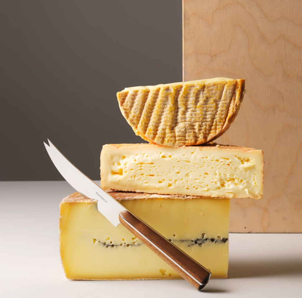 Cheese knife from Couteaux d'orfèvre collection photographed with three different cheeses on a white table in front of a brown background