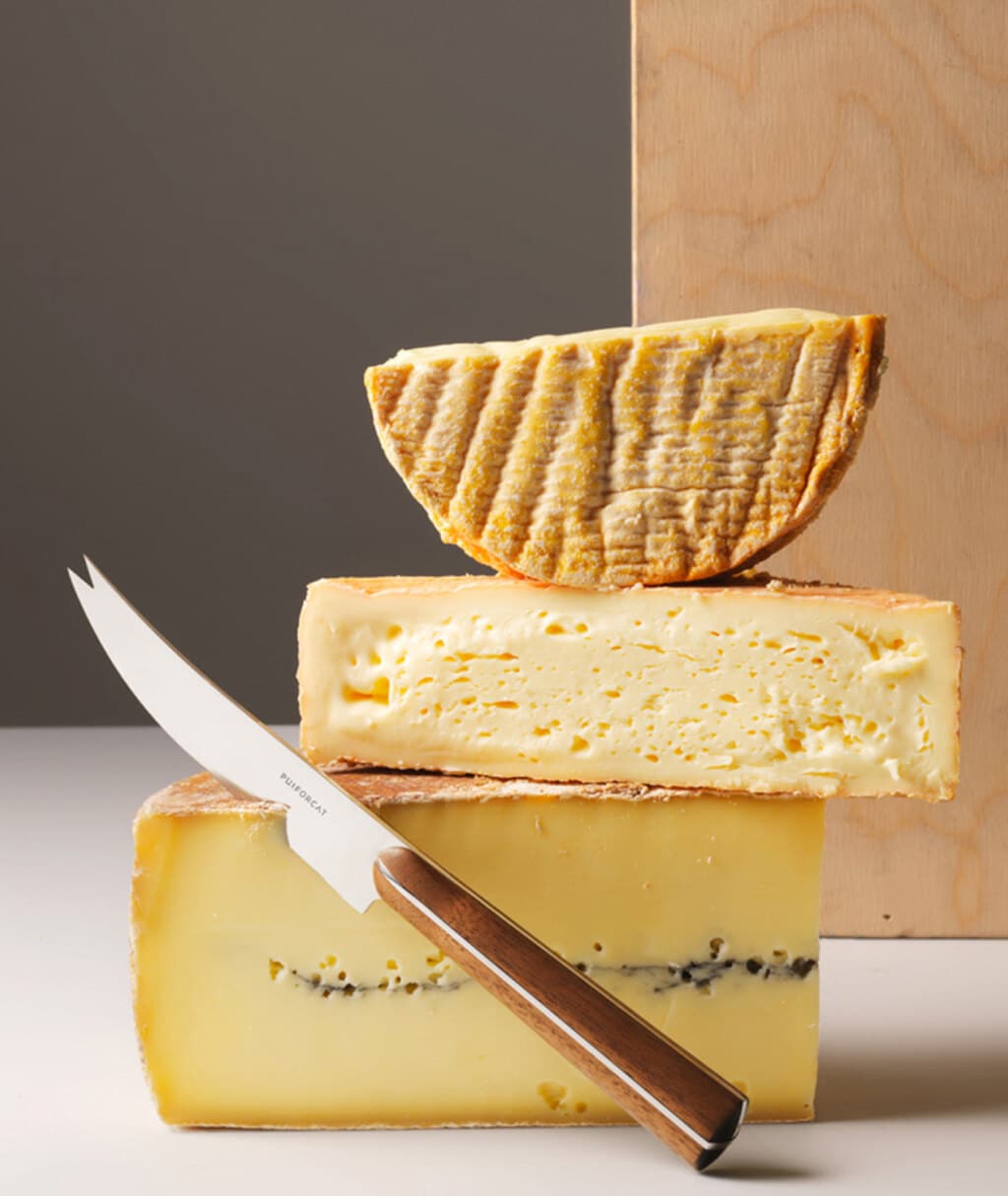 Couteau à fromage de la collection Couteaux d'orfèvre posé à côté d'une pile de trois fromages, photo prise sur un fond en bois